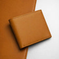 The Saddle bifold wallet in smooth tan finished leather, closed, resting on a leather surface